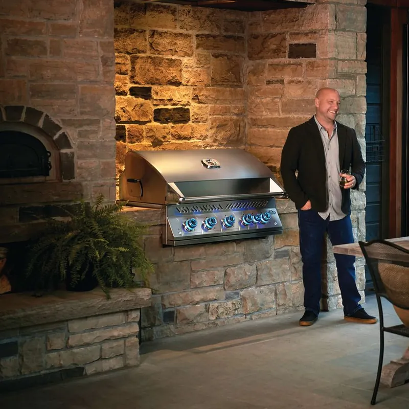 Napoleon Built-in 700 Series gas grill set into the stone wall of an outdoor kitchen beside a wood-fired oven, a host standing next to it at dusk