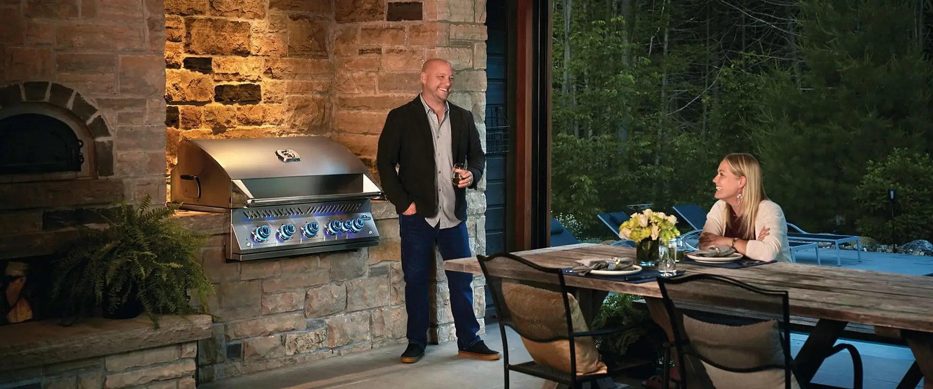 Napoleon Built-in 700 Series gas grill set into the stone wall of an outdoor kitchen beside a wood-fired oven, a host standing next to it at dusk