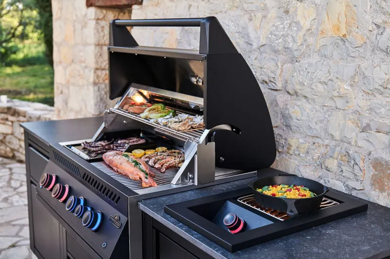 Black Napoleon built-in gas grill with the lid open and a side burner, cooking meat and vegetables in a stone outdoor kitchen