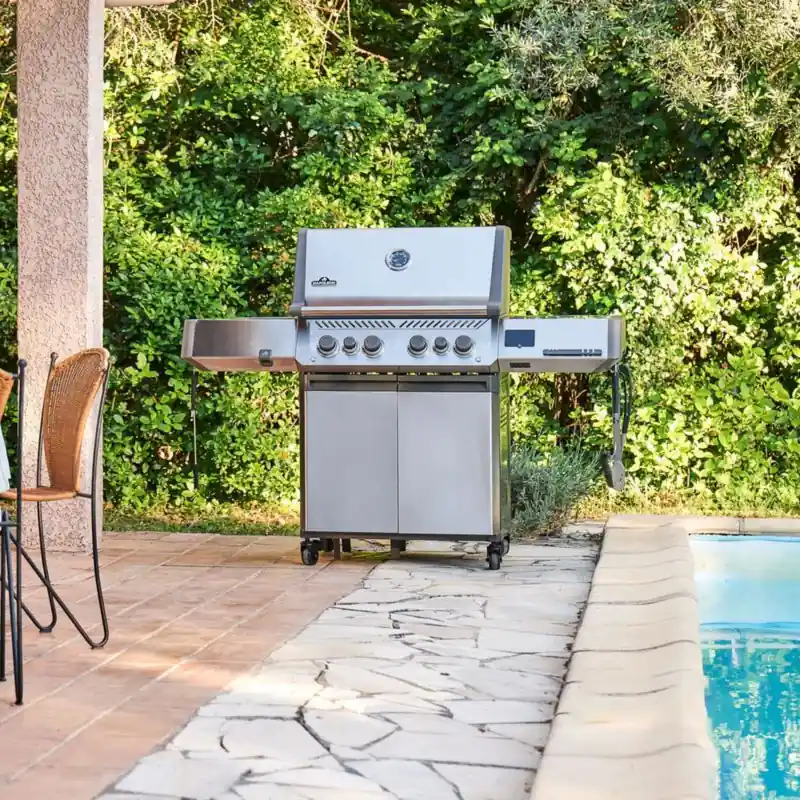 Napoleon Prestige 500 stainless steel gas grill on the terrace of a villa with blue shutters, next to a set table and a pool