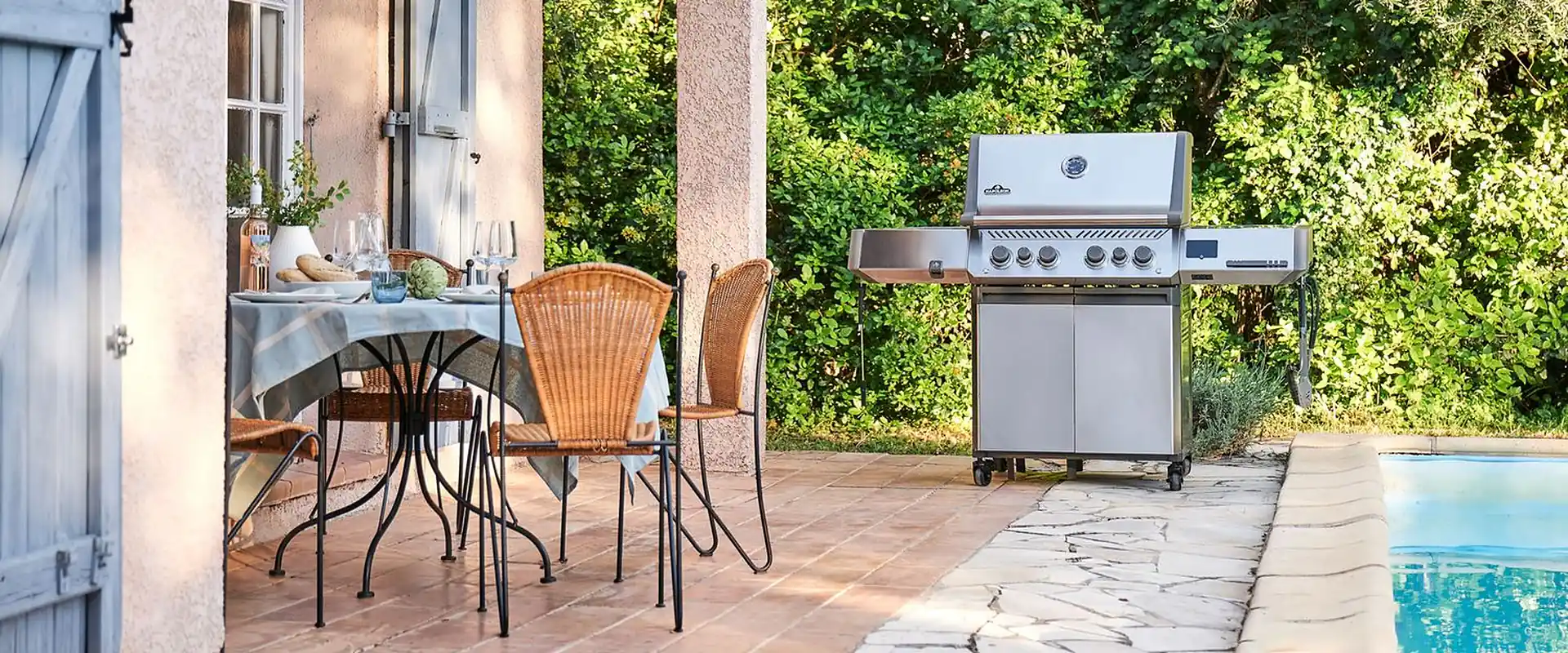 Napoleon Prestige 500 stainless steel gas grill on the terrace of a villa with blue shutters, next to a set table and a pool