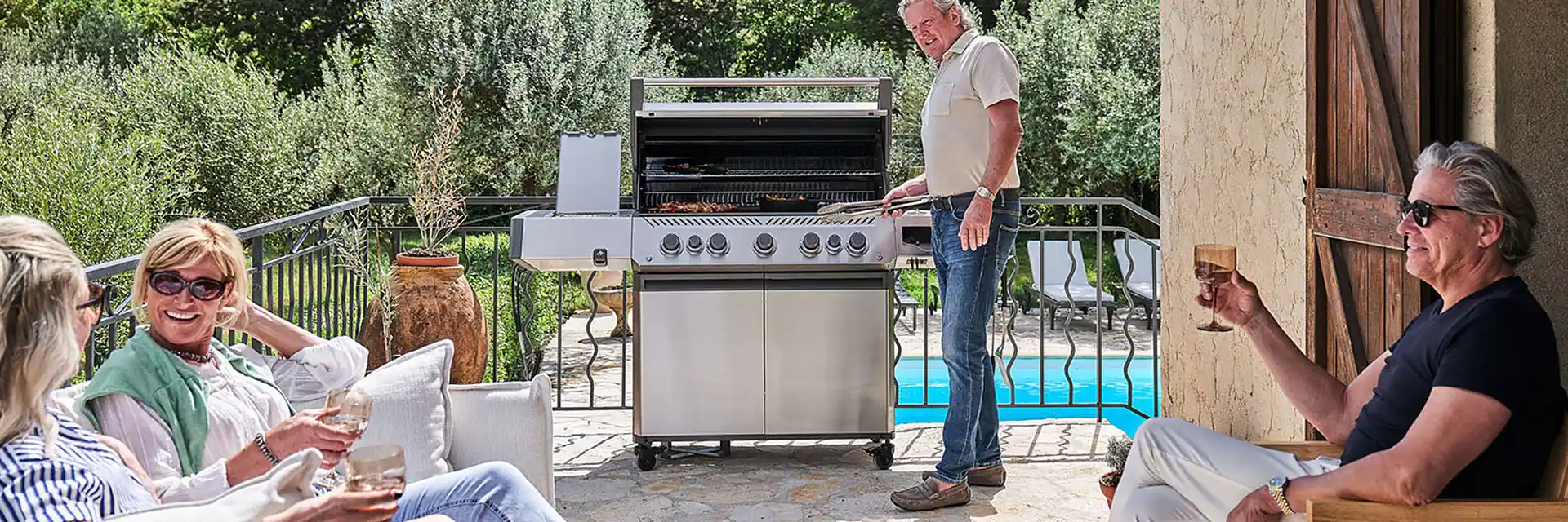 Napoleon Prestige 665 gas barbecue on a stone terrace beside a pool, with a man grilling while friends relax on garden sofas