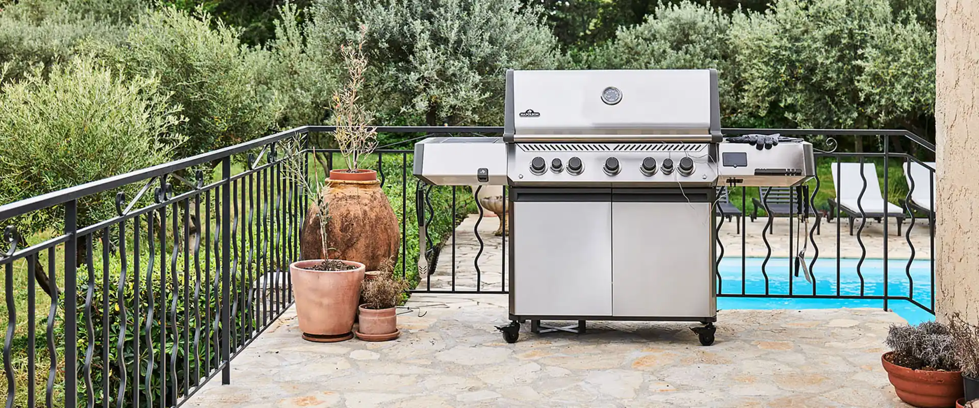 Napoleon Prestige 665 stainless steel gas grill on a stone terrace with terracotta pots, olive trees and a pool behind an iron railing