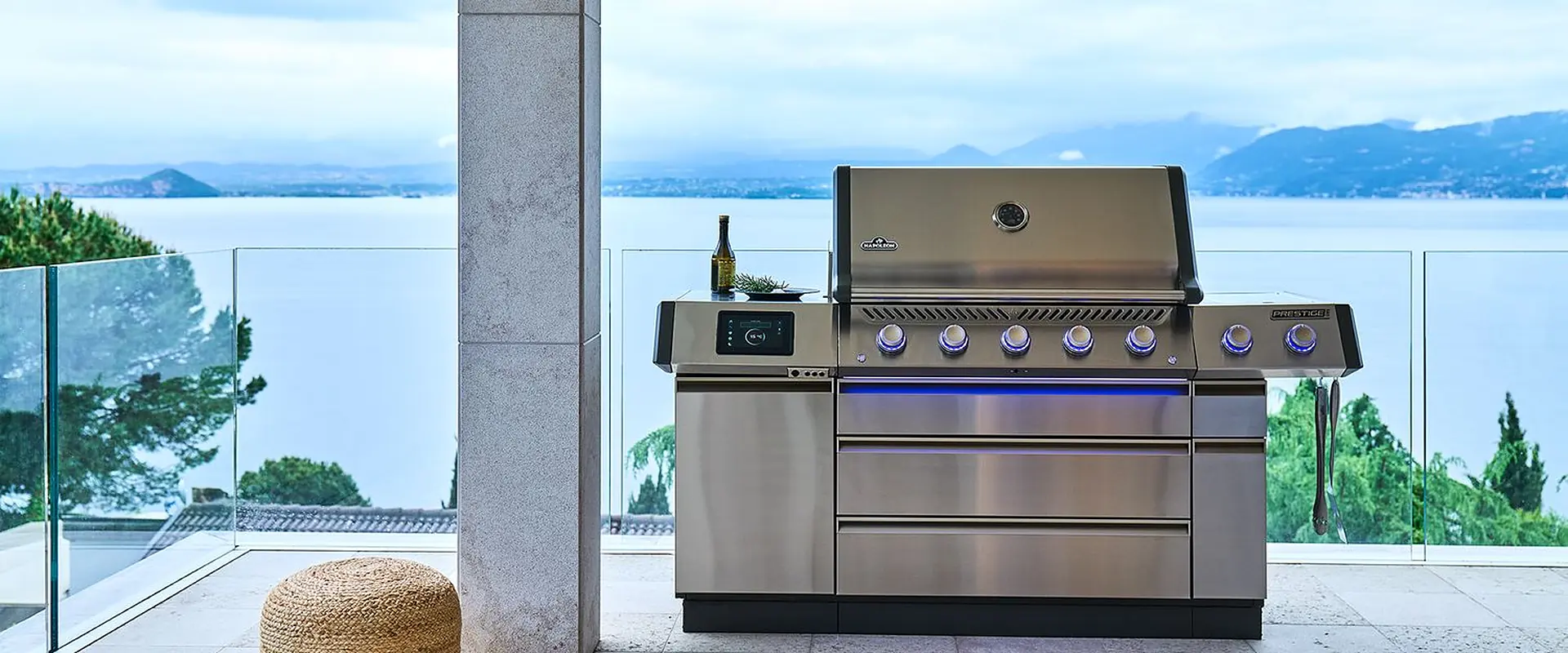 Napoleon Prestige Pro 665 stainless steel gas grill with blue-lit knobs on a covered terrace with a glass balustrade overlooking a lake