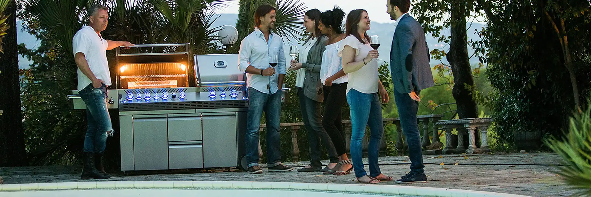 Guests gathered around a lit Napoleon Prestige PRO 825 gas barbecue beside a pool in the evening, under palm trees