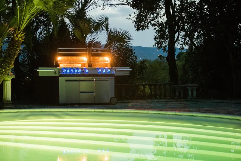 Napoleon Prestige Pro 825 gas grill with its lights on beside a swimming pool at night