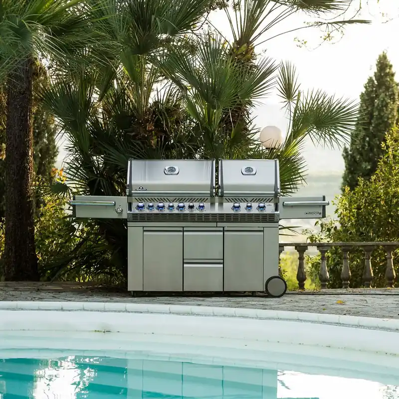 Napoleon Prestige Pro 825 stainless steel gas grill with two lids on a stone terrace between palm trees, beside a pool