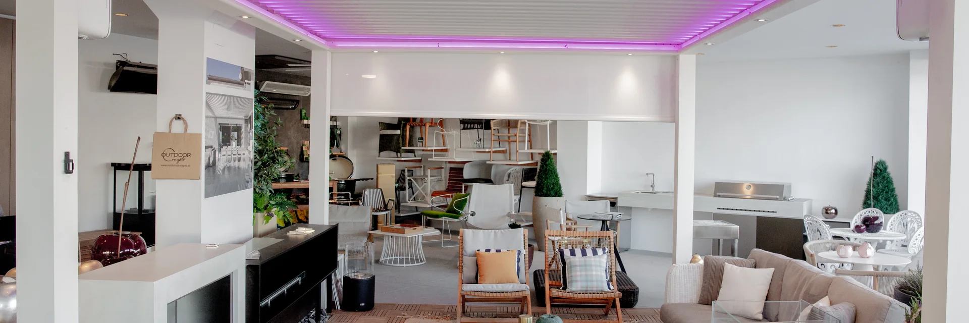 White pergola with LED lighting over a lounge area in the Outdoor Concepts showroom, with outdoor chairs and a kitchen display behind