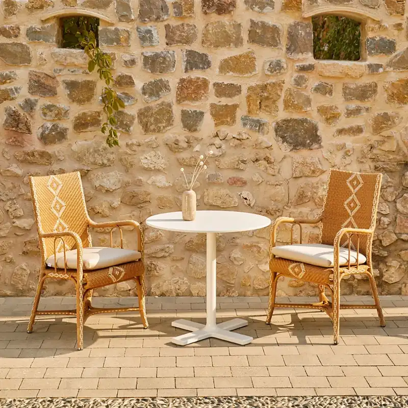 Two Point Charleston woven armchairs at a white bistro table on terracotta tiles, in front of an old stone wall