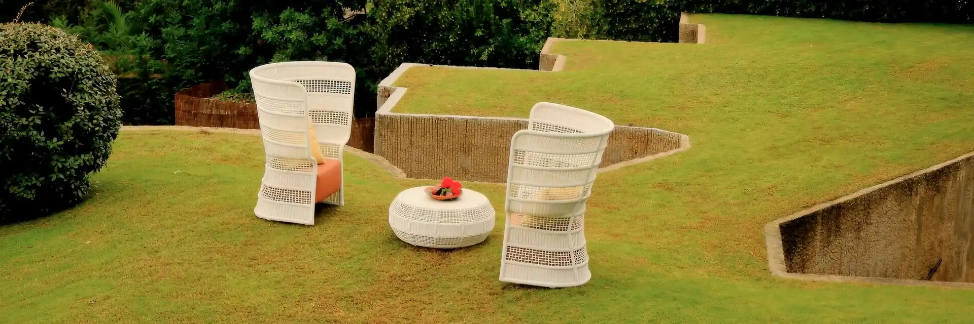 Point Escala woven armchairs and a round pouf on a stepped green lawn, with trees behind