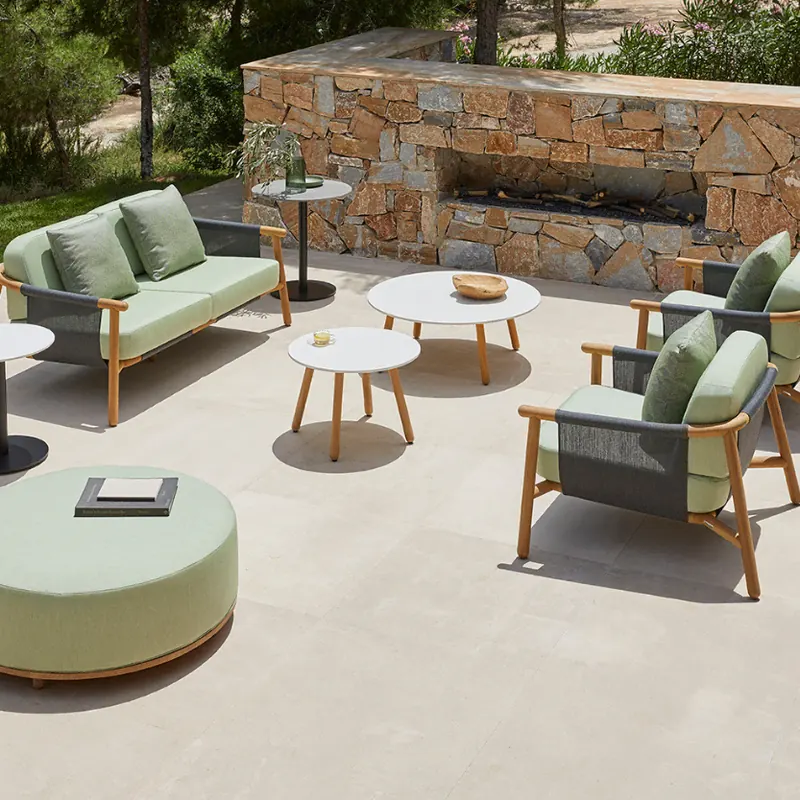 Point Hamp teak sofas with green cushions, a round pouf and side tables on a pale terrace beside a stone wall