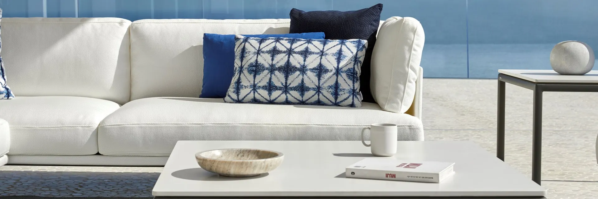 Close view of a white Point Long Island sofa with blue patterned cushions beside a low table, on a terrace above the sea
