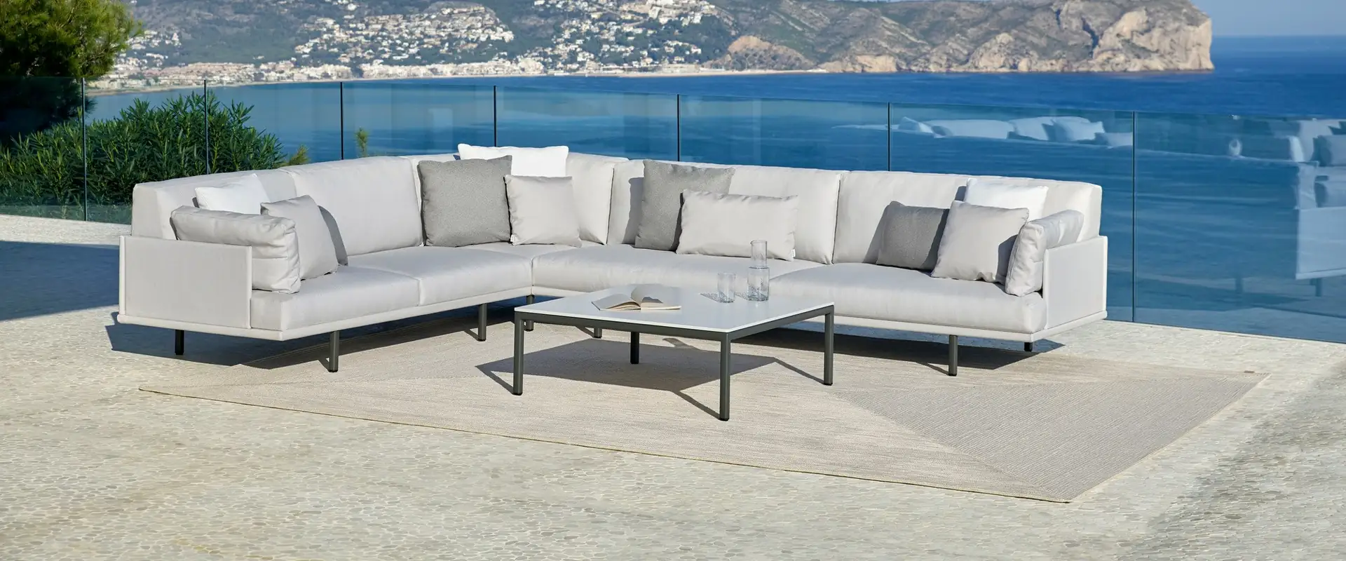 Point Long Island corner sofa in white on a terrace with a glass balustrade, overlooking the sea and a rocky headland