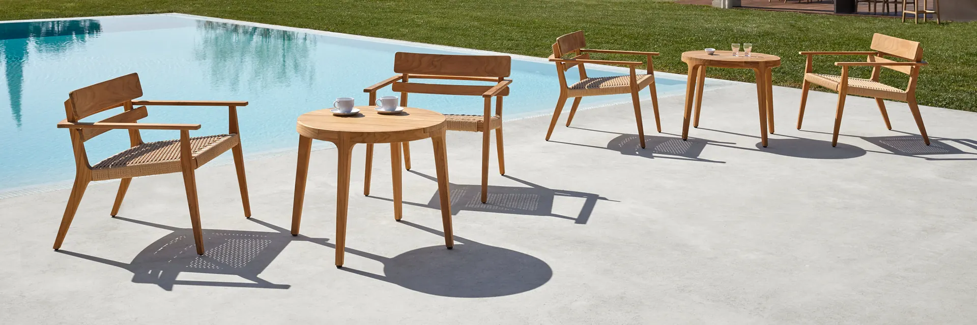 Point Paralel teak armchairs and side tables on a terrace beside a pool, with a lawn and trees behind