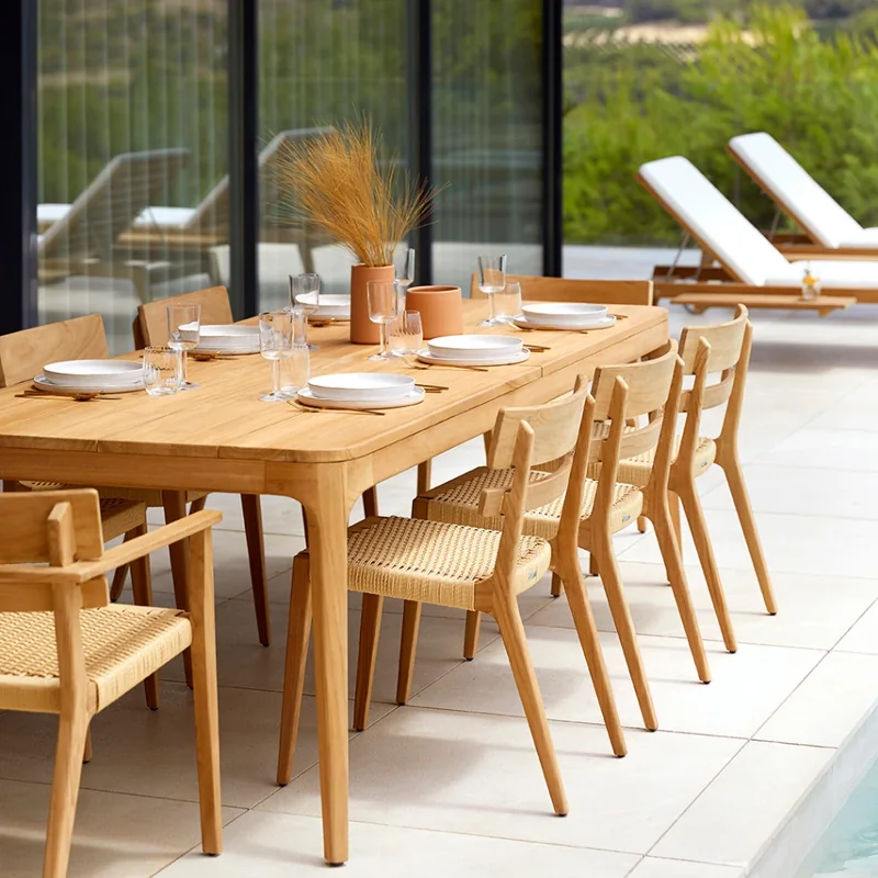 Point Paralel teak dining table set for a meal with teak armchairs on a pale terrace beside a pool