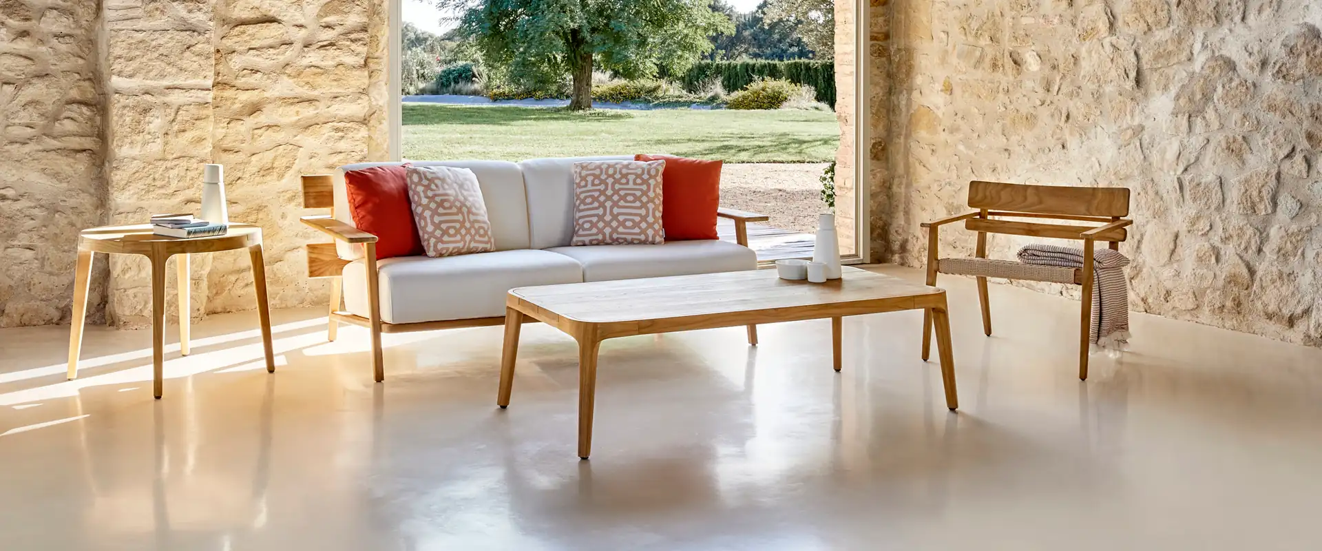 Point Paralel teak sofa, armchair and low tables in a stone-walled porch with a large arch opening onto a garden