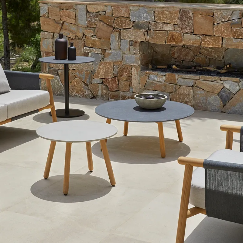 Point Round coffee and side tables with teak legs on a pale stone terrace in front of a dry-stone wall