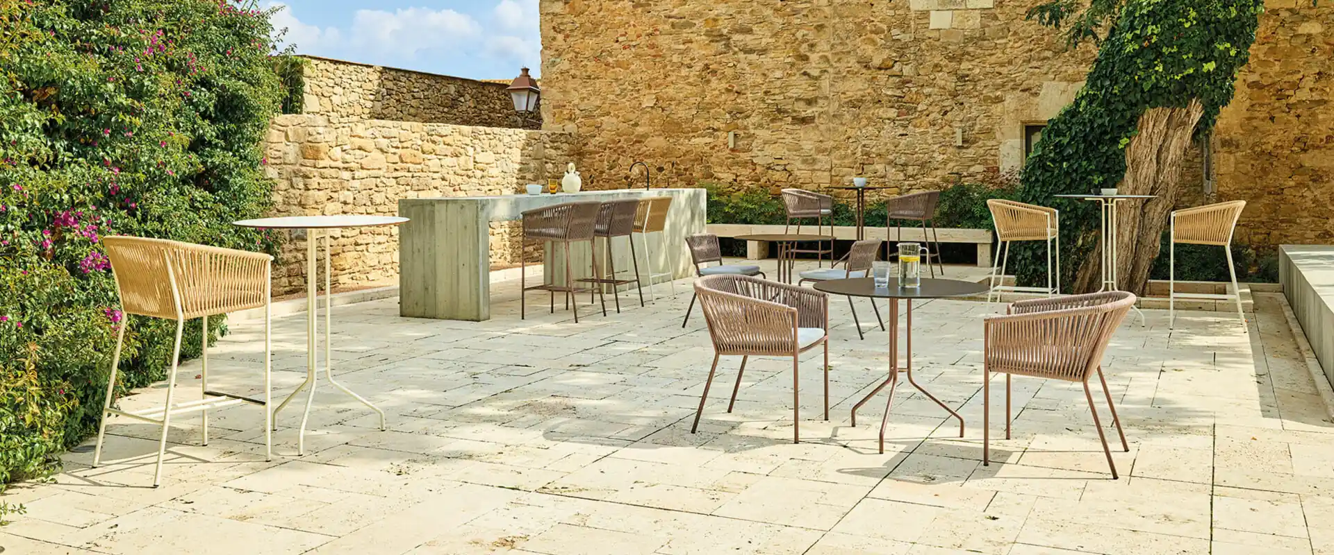 Point Weave rope chairs and round tables on the paved terrace of an old stone house, with flowering shrubs