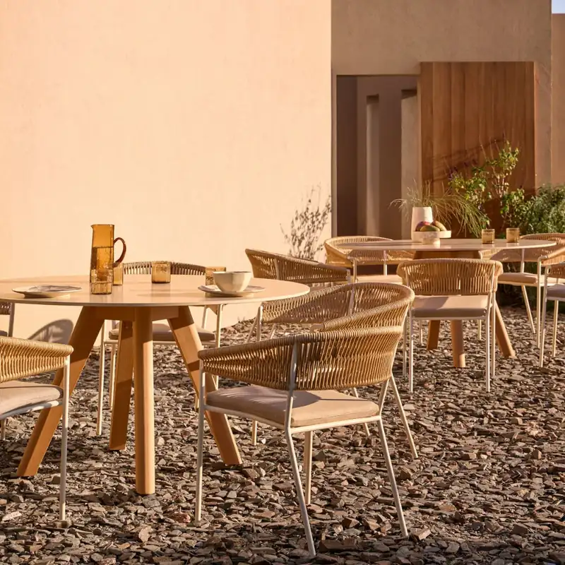 Point Weave rope dining chairs around a round teak table on a sunny terrace beside a terracotta-colored house
