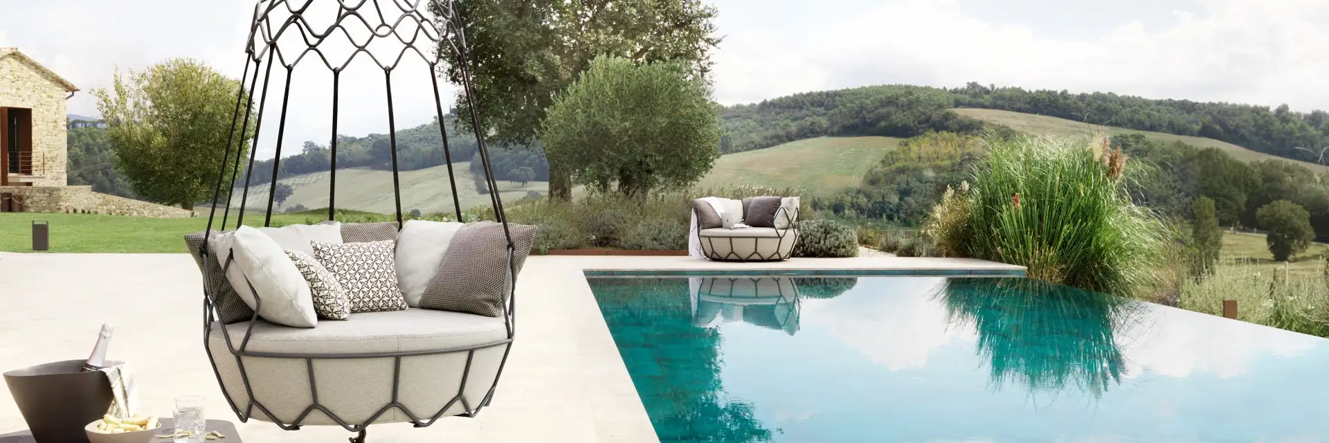 Roberti Gravity hanging swing sofa with a graphite frame and pale cushions, suspended beside a pool with rolling hills behind