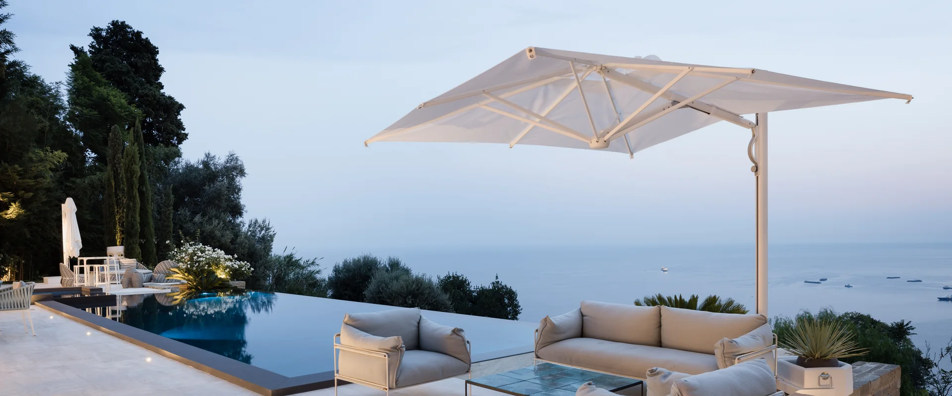 White Scolaro Astro cantilever parasol shading a lounge set beside an infinity pool on a villa terrace overlooking the sea at dusk, with cypresses on the hillside