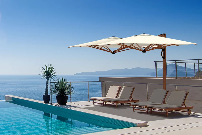 Scolaro Dual V parasol with two ecru canopies on a timber mast over wooden sun loungers beside an infinity pool, overlooking the sea