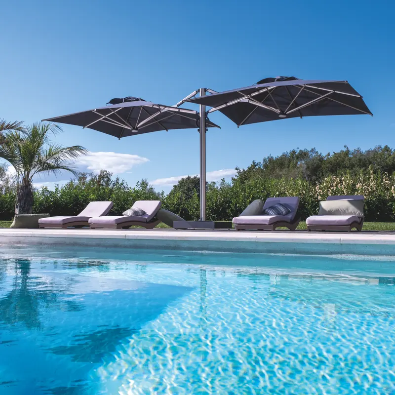 Dark Scolaro Galaxia Dual T parasol with two canopies on a single mast shading sun loungers at the edge of a turquoise pool, with palm trees and a hedge behind