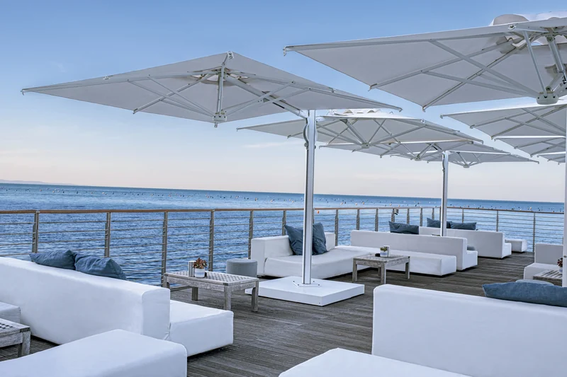 White Scolaro Galaxia Dual T parasols with twin canopies on white masts over white lounge sofas on a wooden deck beside the sea