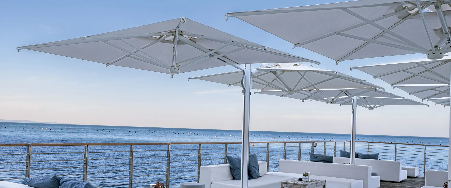 White Scolaro Galaxia Dual T parasols with twin canopies on white masts over white lounge sofas on a wooden deck beside the sea