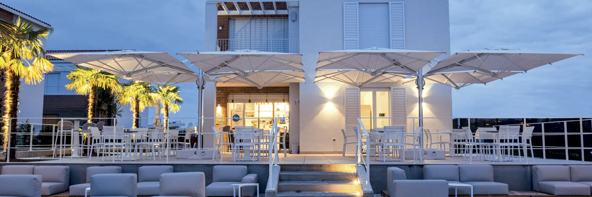 White four-canopy Scolaro Galaxia Quattro parasols over the lounge terrace of a white clubhouse at dusk, with palms and sofas