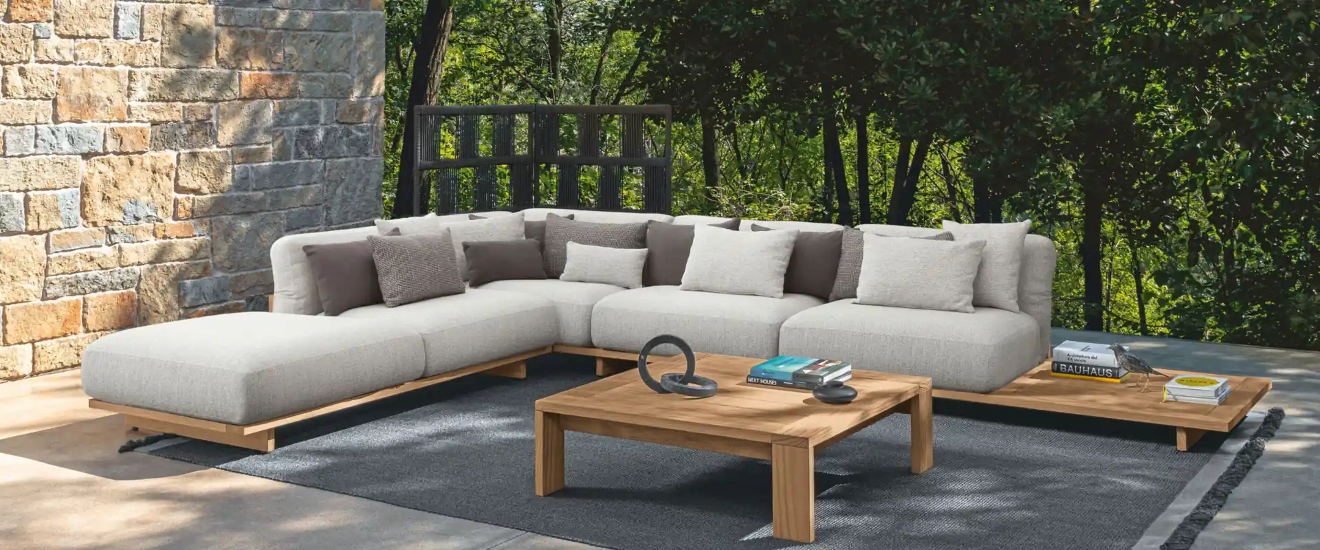 Talenti Argo sectional sofa with a teak base and pale cushions on a terrace beside a stone wall, surrounded by trees