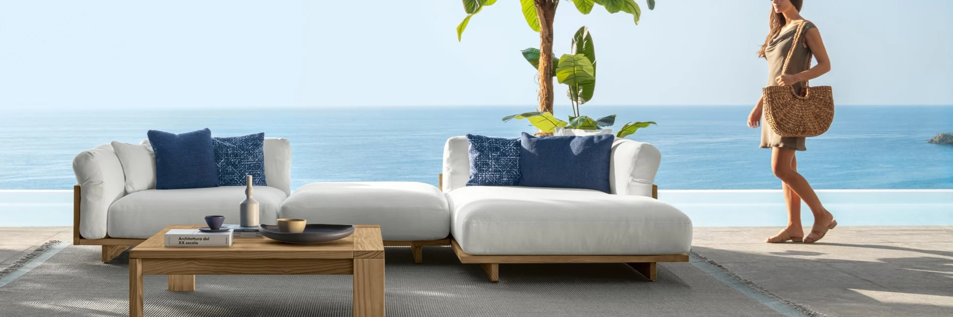 Talenti Argo sofa with navy cushions and a teak coffee table on a seafront terrace, with a woman walking past a banana plant