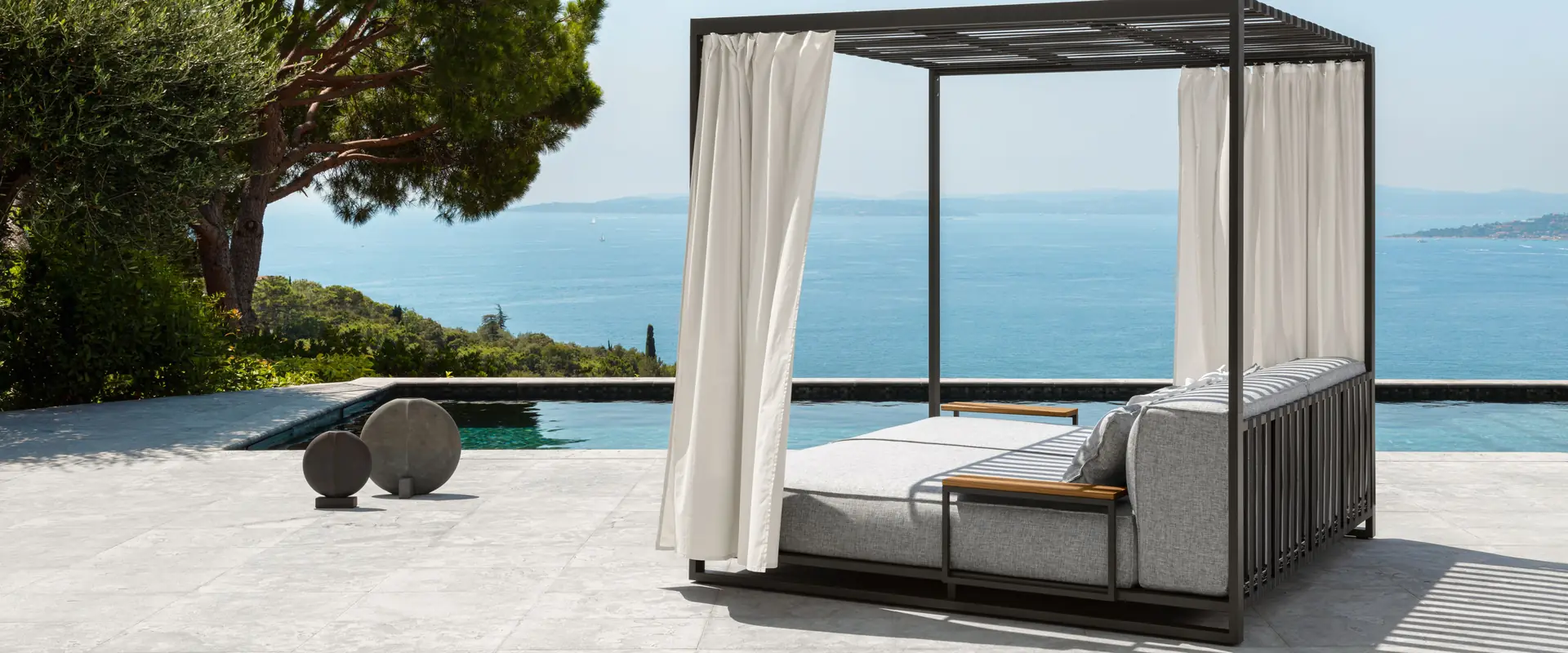 Talenti Casilda daybed with a black canopy frame and white curtains on a terrace by the sea, beside a pine tree