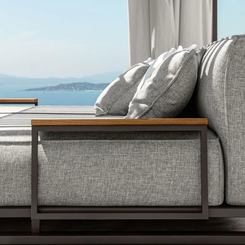 Close view of a Talenti Casilda sofa with gray cushions and a wooden armrest shelf, the sea in the background