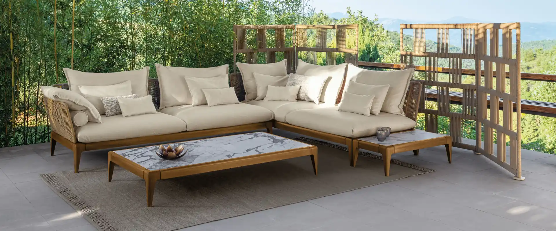 Talenti Cruise teak corner sofa with pale cushions and a coffee table on a stone terrace bordered by greenery