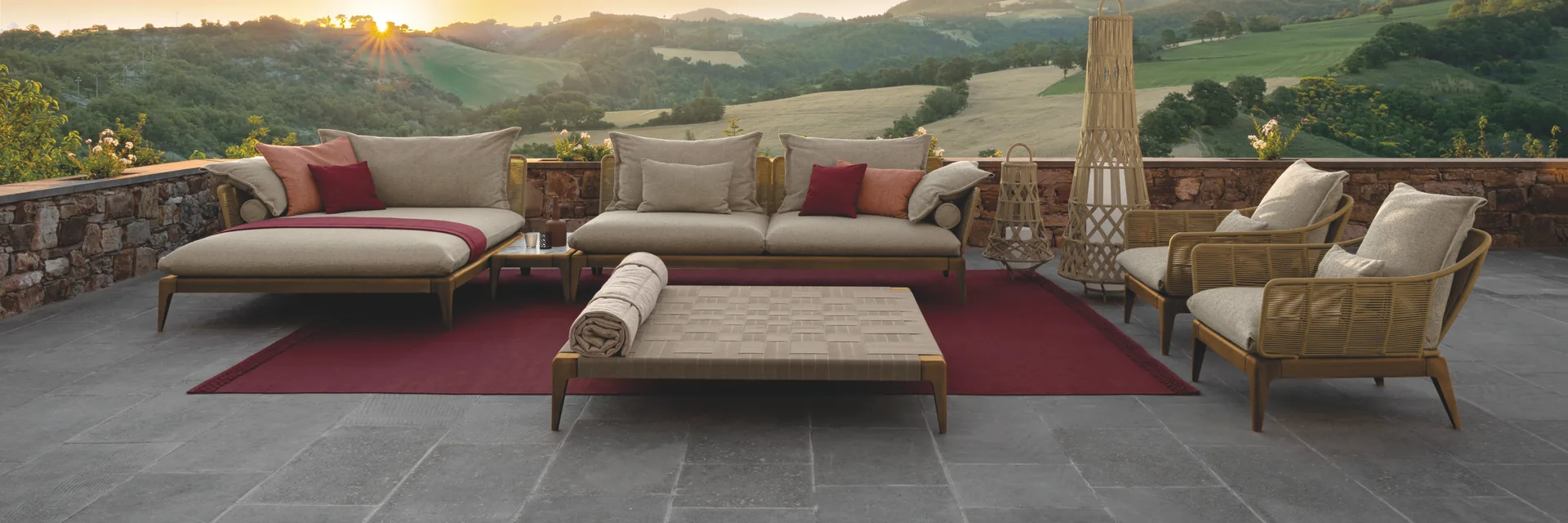 Talenti Cruise teak sofas and daybeds on a terrace with a red rug, facing green hills under a sunset sky