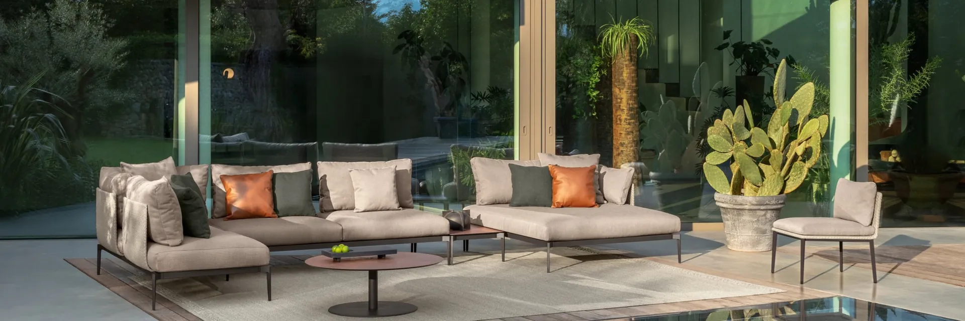 Talenti Leaf sofa and armchairs with orange cushions on the terrace of a glass-fronted house, reflected in the pool