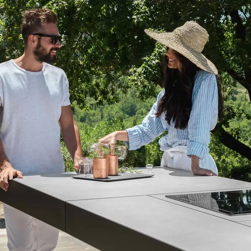 Couple preparing drinks at a Talenti outdoor kitchen island with an induction hob and sink, under trees on a hillside terrace