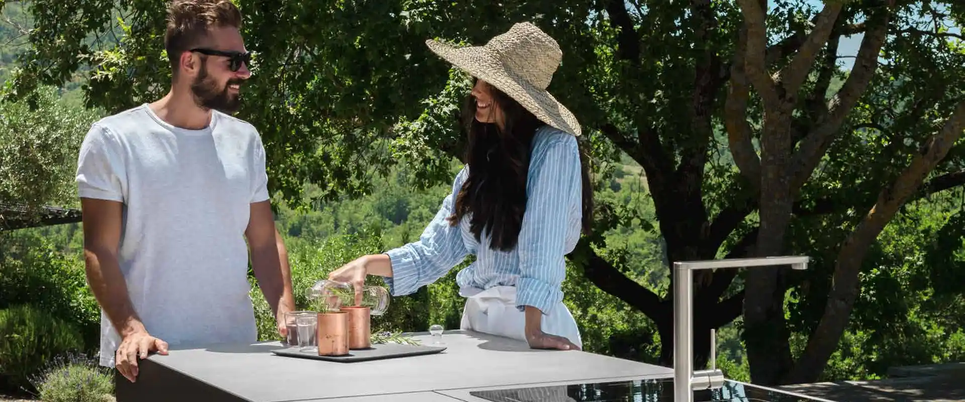 Couple preparing drinks at a Talenti outdoor kitchen island with an induction hob and sink, under trees on a hillside terrace