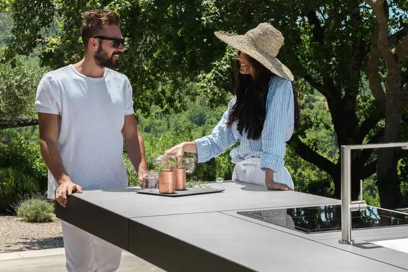 Couple preparing drinks at a Talenti outdoor kitchen island with an induction hob and sink, under trees on a hillside terrace