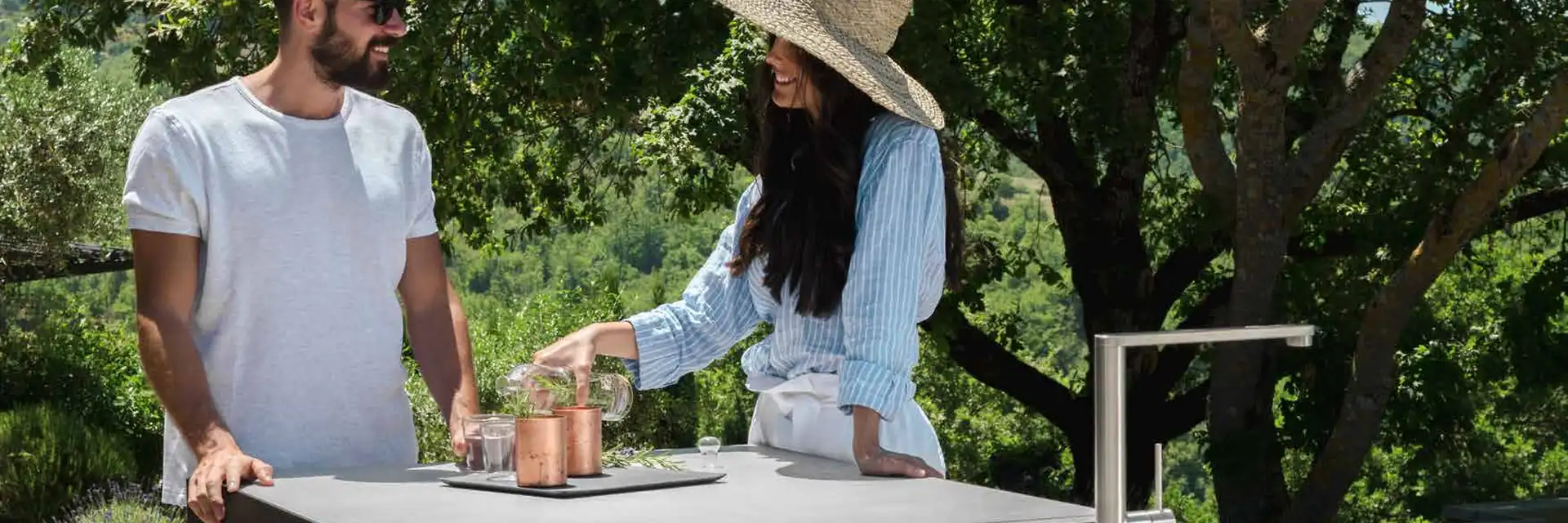 Couple preparing drinks at a Talenti outdoor kitchen island with an induction hob and sink, under trees on a hillside terrace
