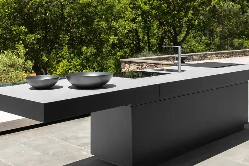 Dark Talenti outdoor kitchen island with a floating worktop, woks and a tap on a stone pool terrace surrounded by trees