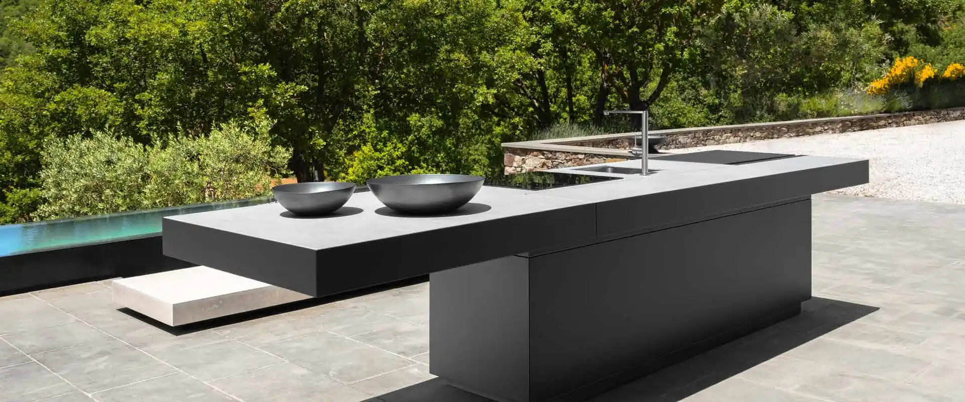 Dark Talenti outdoor kitchen island with a floating worktop, woks and a tap on a stone pool terrace surrounded by trees