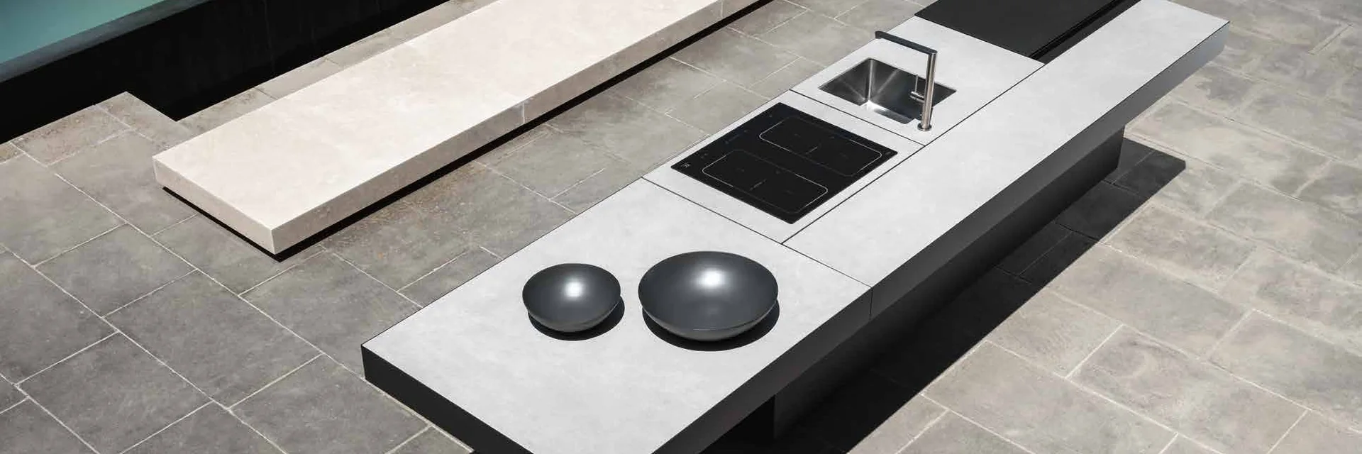 Talenti outdoor kitchen island with an induction hob, a sink and woks on a stone terrace beside a reflecting pool, seen from above