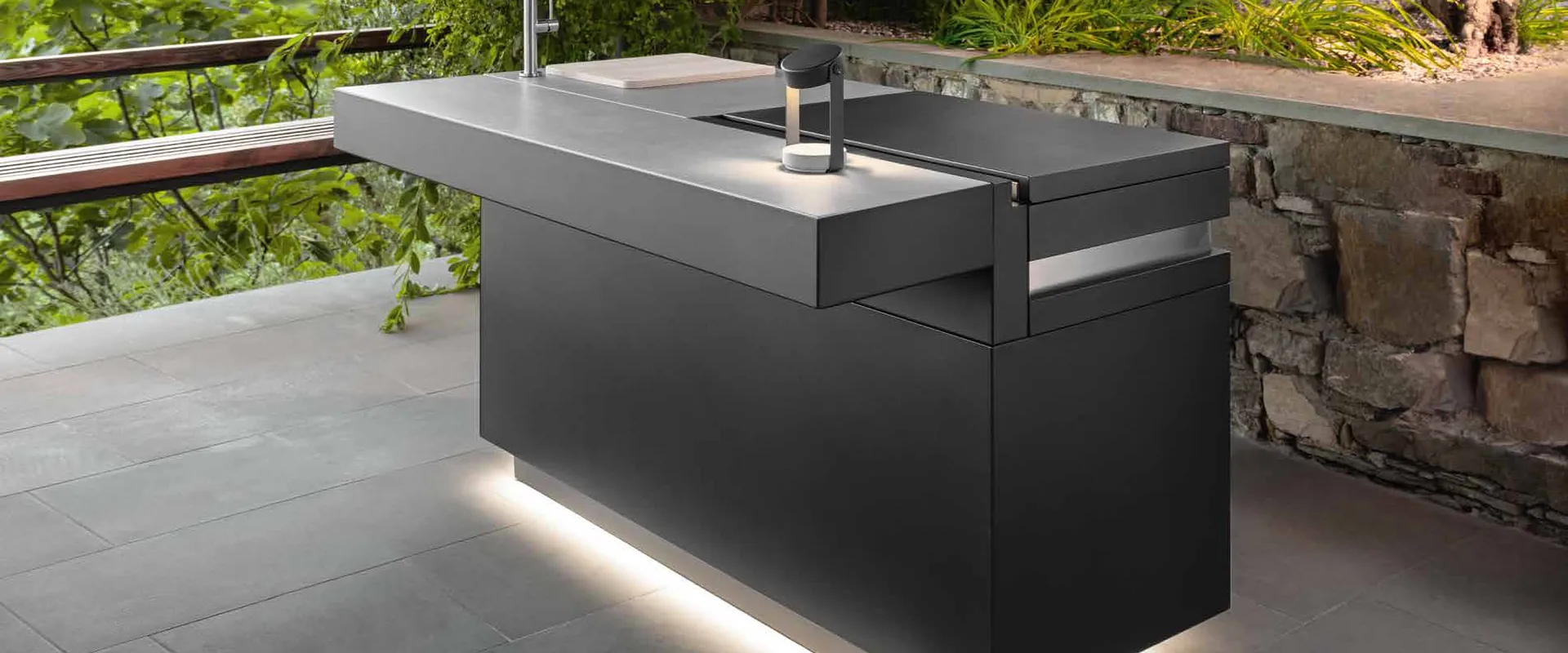 Dark Talenti outdoor kitchen island with a lit plinth on a stone terrace with a dry-stone wall and Mediterranean planting