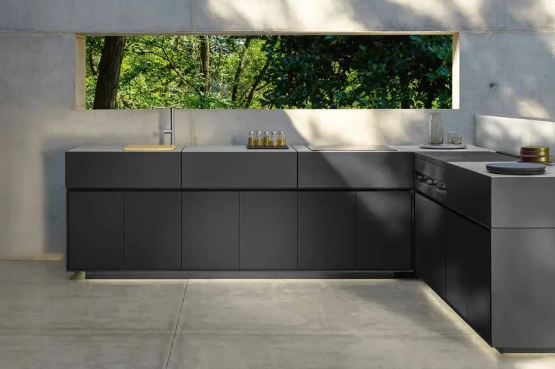 Dark L-shaped Talenti outdoor kitchen with a lit plinth in a concrete courtyard with a window onto the garden