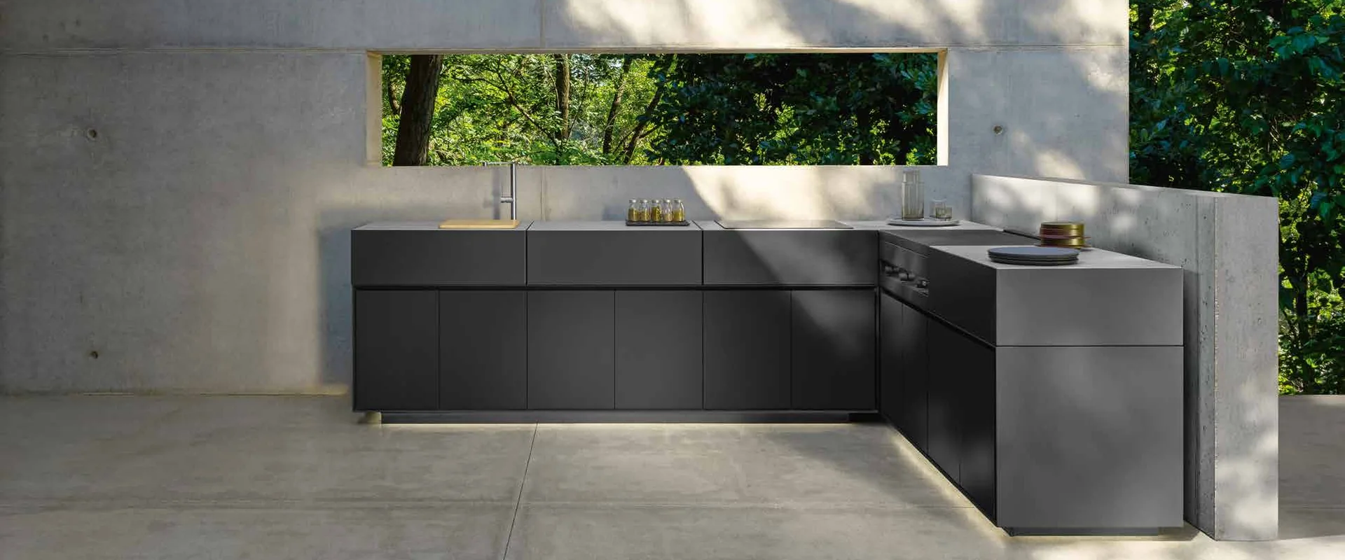 Dark L-shaped Talenti outdoor kitchen with a lit plinth in a concrete courtyard with a window onto the garden