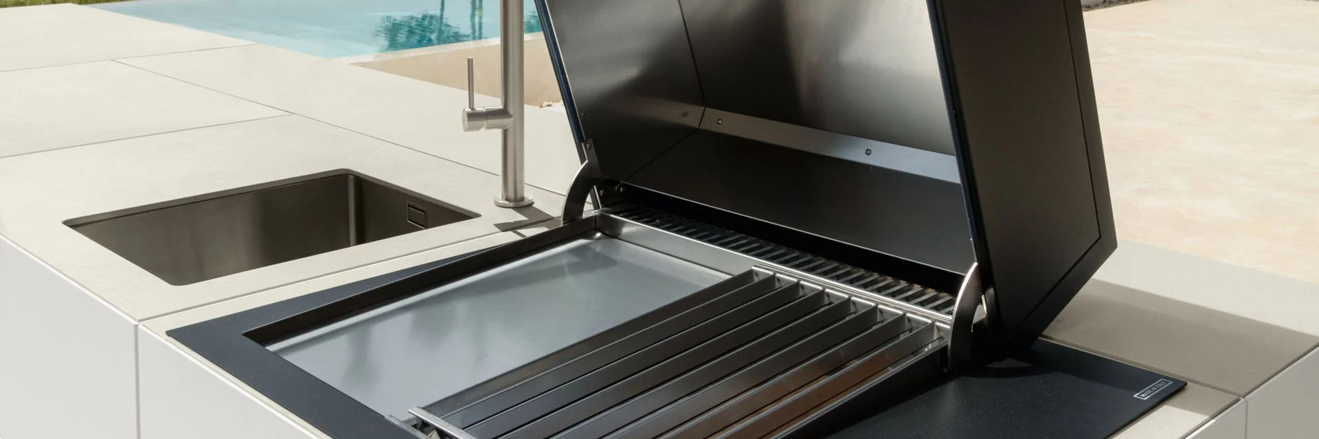 Built-in grill with its lid open in a white Talenti Tikal outdoor kitchen island, with a lawn and trees behind