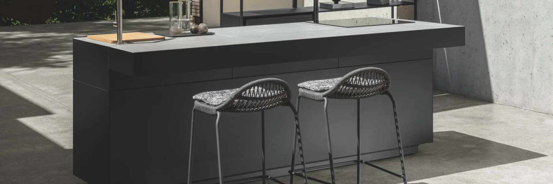 Dark Talenti Tikal outdoor kitchen island with two bar stools in a concrete courtyard with open shelving