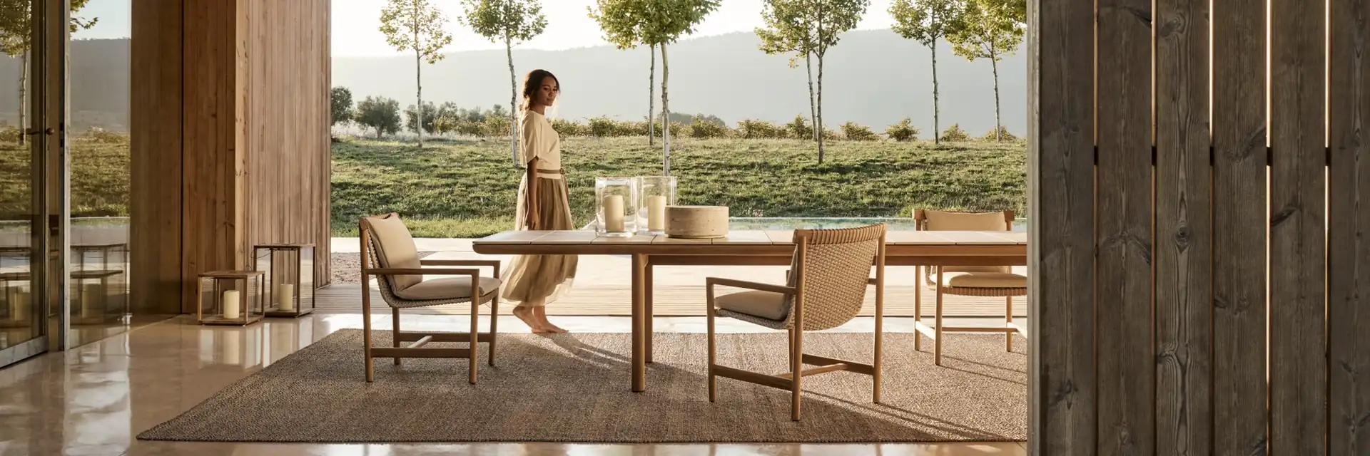 Tribù Amanu dining table and chairs on a terrace seen through a timber pavilion, with a woman walking past and trees beyond
